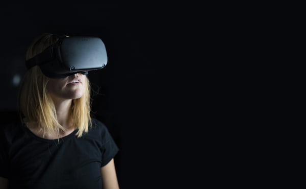 Uma mulher loira com camiseta preta com um par de óculos VR em uma sala escura.