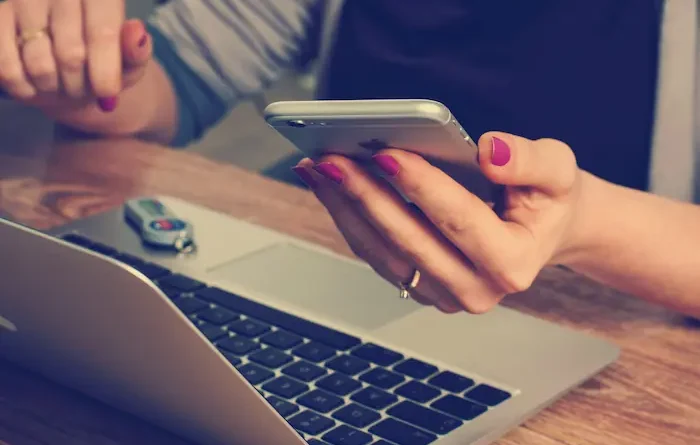 Uma mulher com o celular na mão mexe no notebook que está em cima de uma mesa.